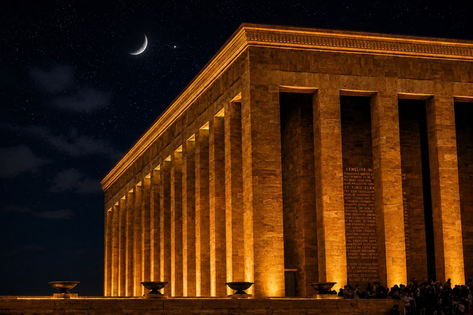 Anıtkabir's colonnade lit gold at night, under a crescent moon.